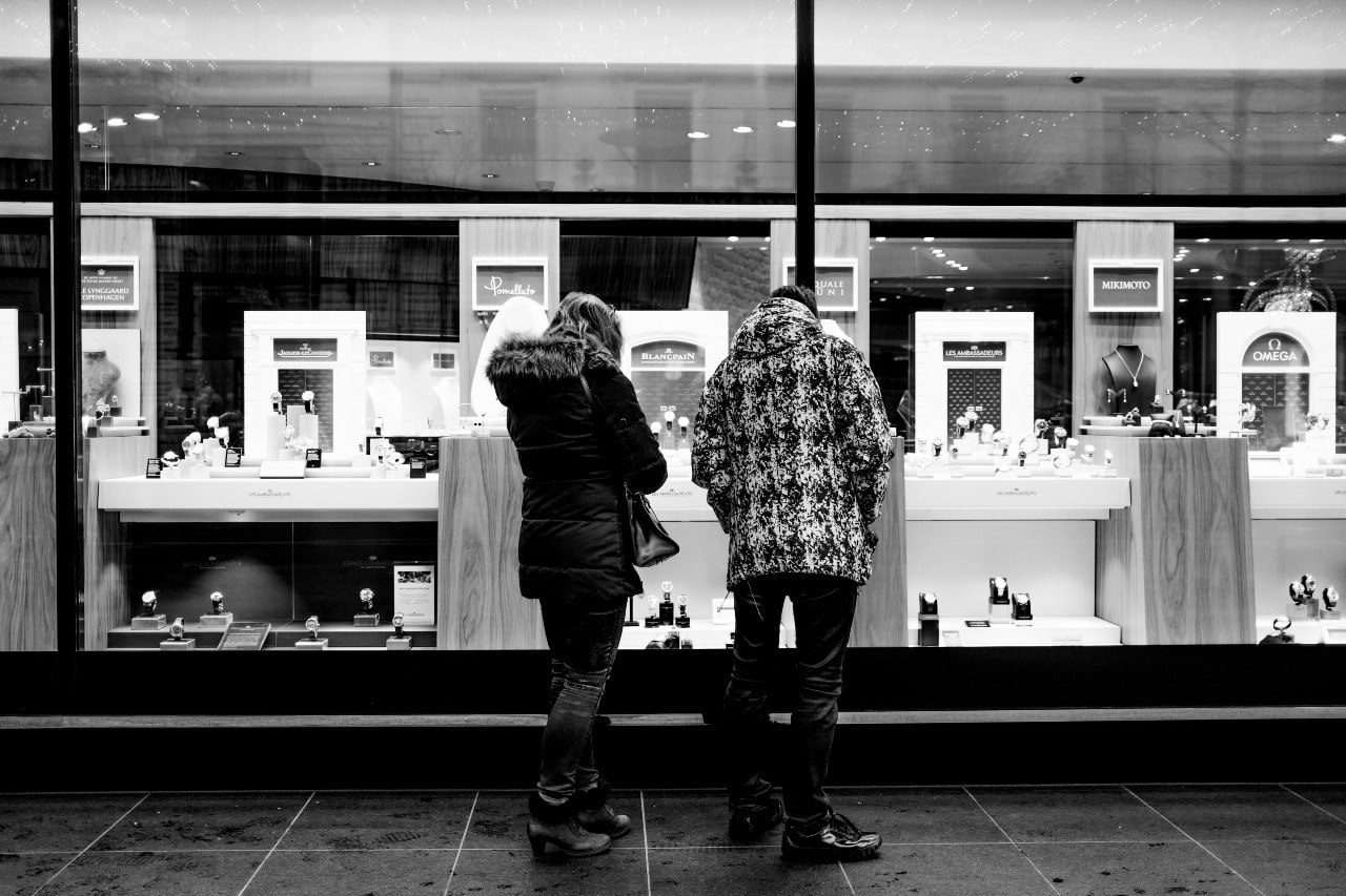 A couple window-shops outside of a closed jewelry store in the city at night