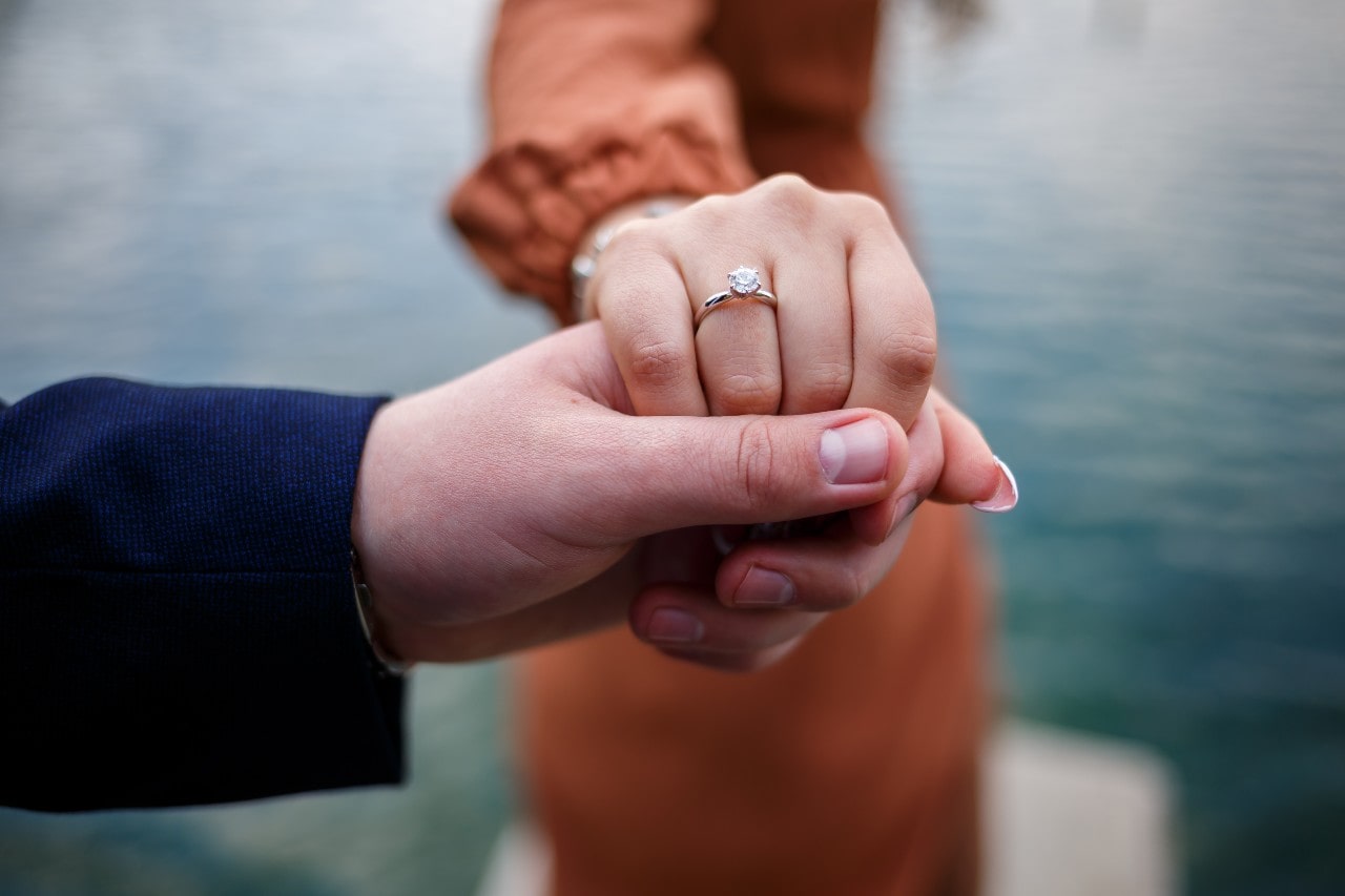 A close-up of a man holding a woman's hand with a solitaire engagement ring