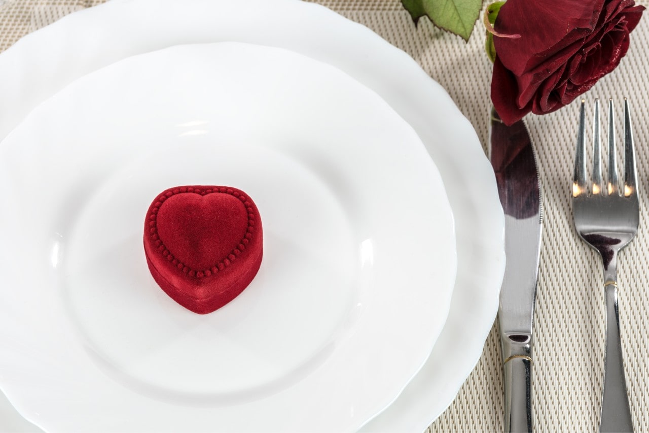 A small heart shape jewelry box on a white plate sitting next to a knife, fork, and single red rose on a table