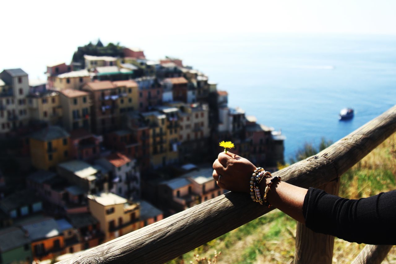 A woman wearing multiple beaded bracelets holds a dandelion while looking over a Greek village on the coast