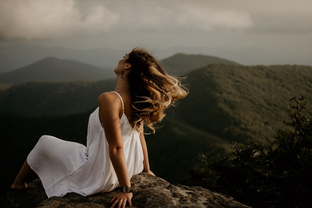 A woman wearing a white sundress and gemstone earrings enjoys the view of mountainous landscapes in Scotland