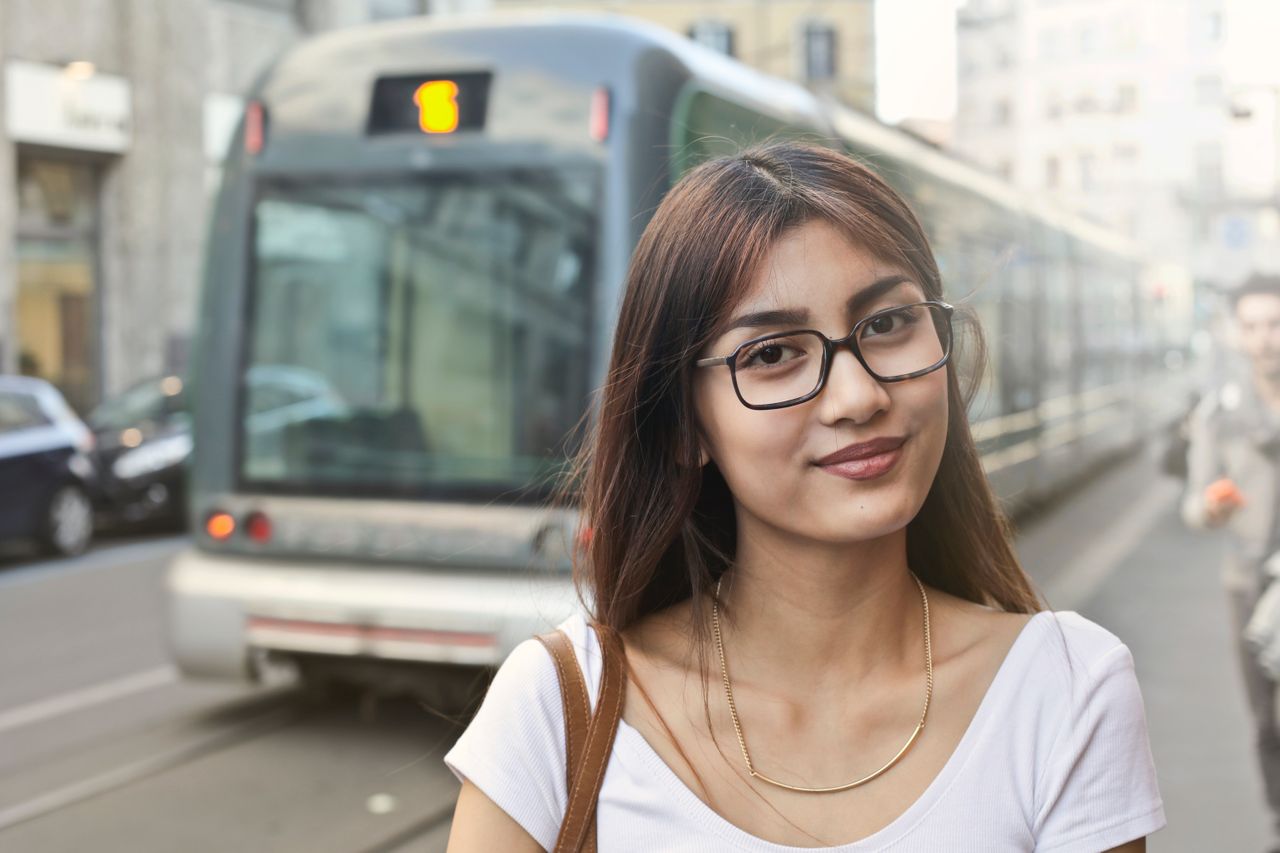 A woman wearing a quirky necklace and glasses stands on a sidewalk in a downtown city in Michigan