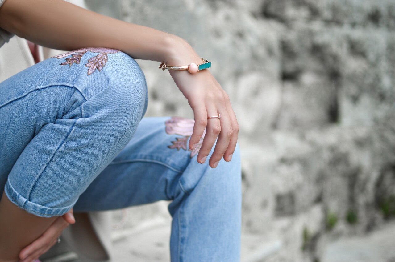 A woman with a gemstone bracelet and embroidered jeans sits against a stone wall