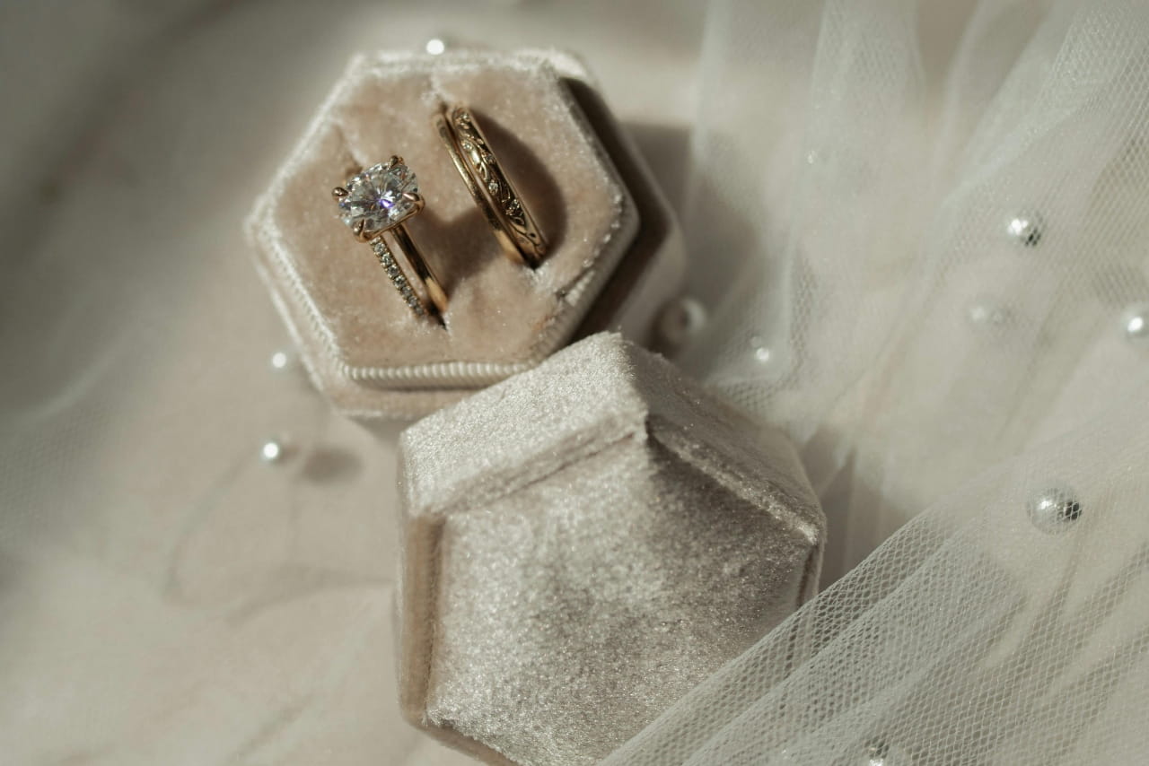 Yellow gold diamond solitaire engagement ring and wedding band, rest inside a soft velvet ring box on sheer fabric decorated with pearls.