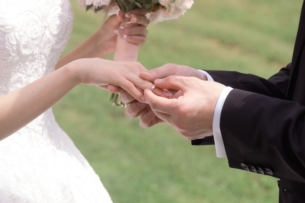 A groom in a black suit gently places a wedding band on the bride's finger as she holds her bouquet.
