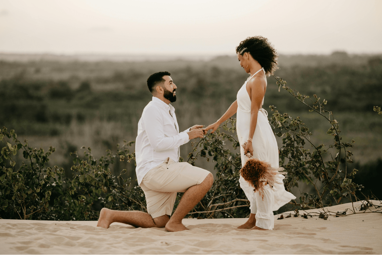 A man kneels on one knee proposing to a woman holding flowers on a sandy landscape.