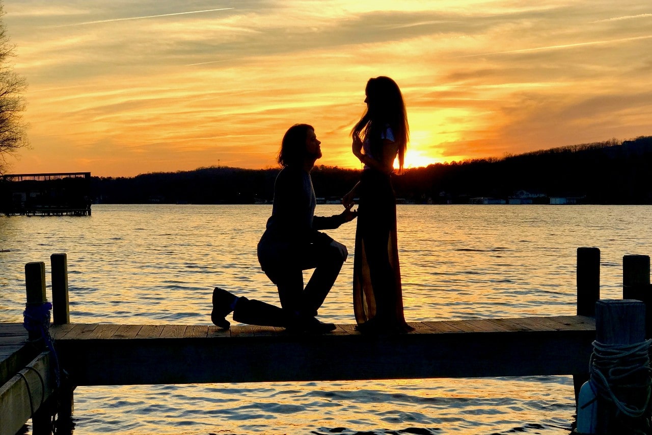 A person kneels on a dock, proposing to another, silhouetted by a golden sunset over a tranquil lake.