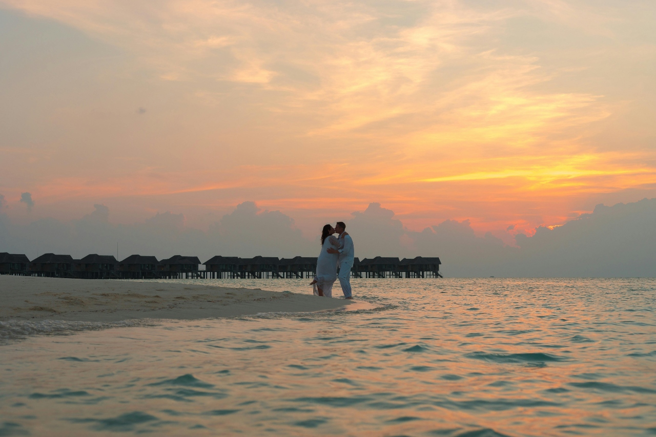 A couple embraces on a beach at sunset, with serene, calm water in the foreground.