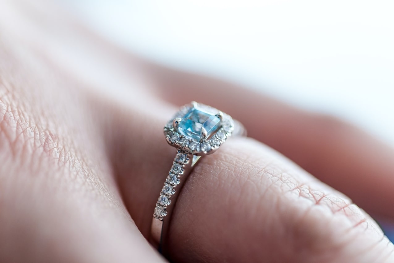 close up image of an engagement ring on someone’s finger that features a halo set blue topaz