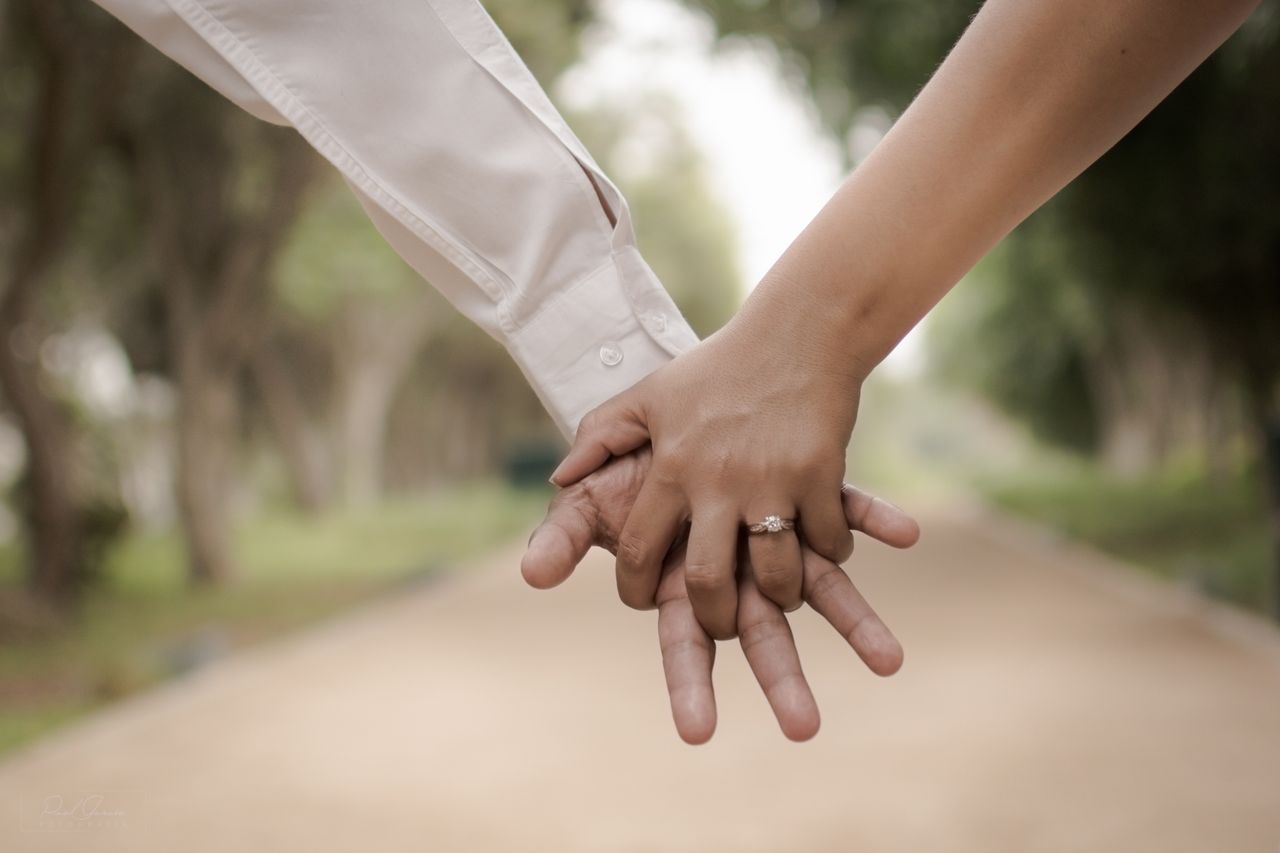 A couple holding hands in front of a trail of greenery, showcasing the woman&rsquo;s beautiful engagement ring.