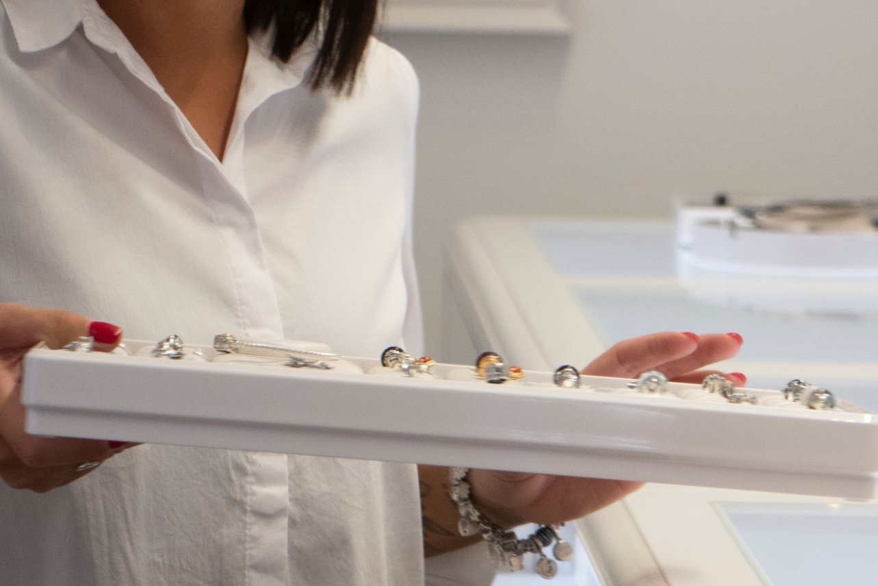 A jewelry store employee wearing a white shirt holds a long white display tray filled with various engagement rings and small charms.