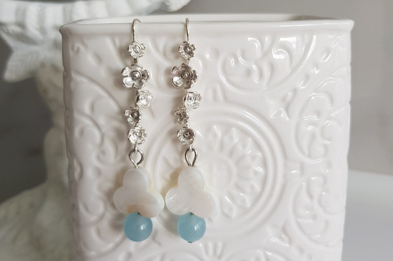 A close-up of silver floral drop earrings hooked on a white porcelain vase, the bottom of the earrings beaded.