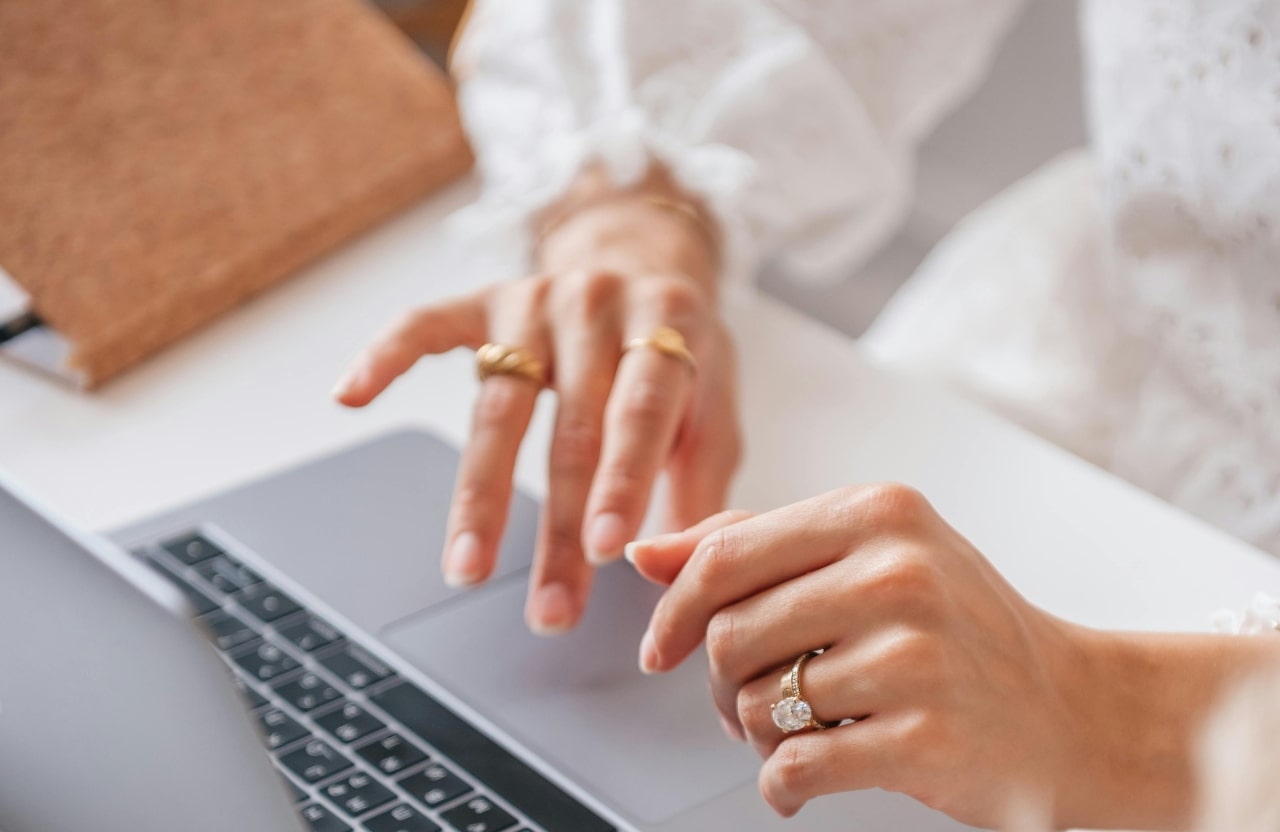 An angled view of a woman typing on a laptop, with two yellow gold bands on one hand and a diamond solitaire on the other.