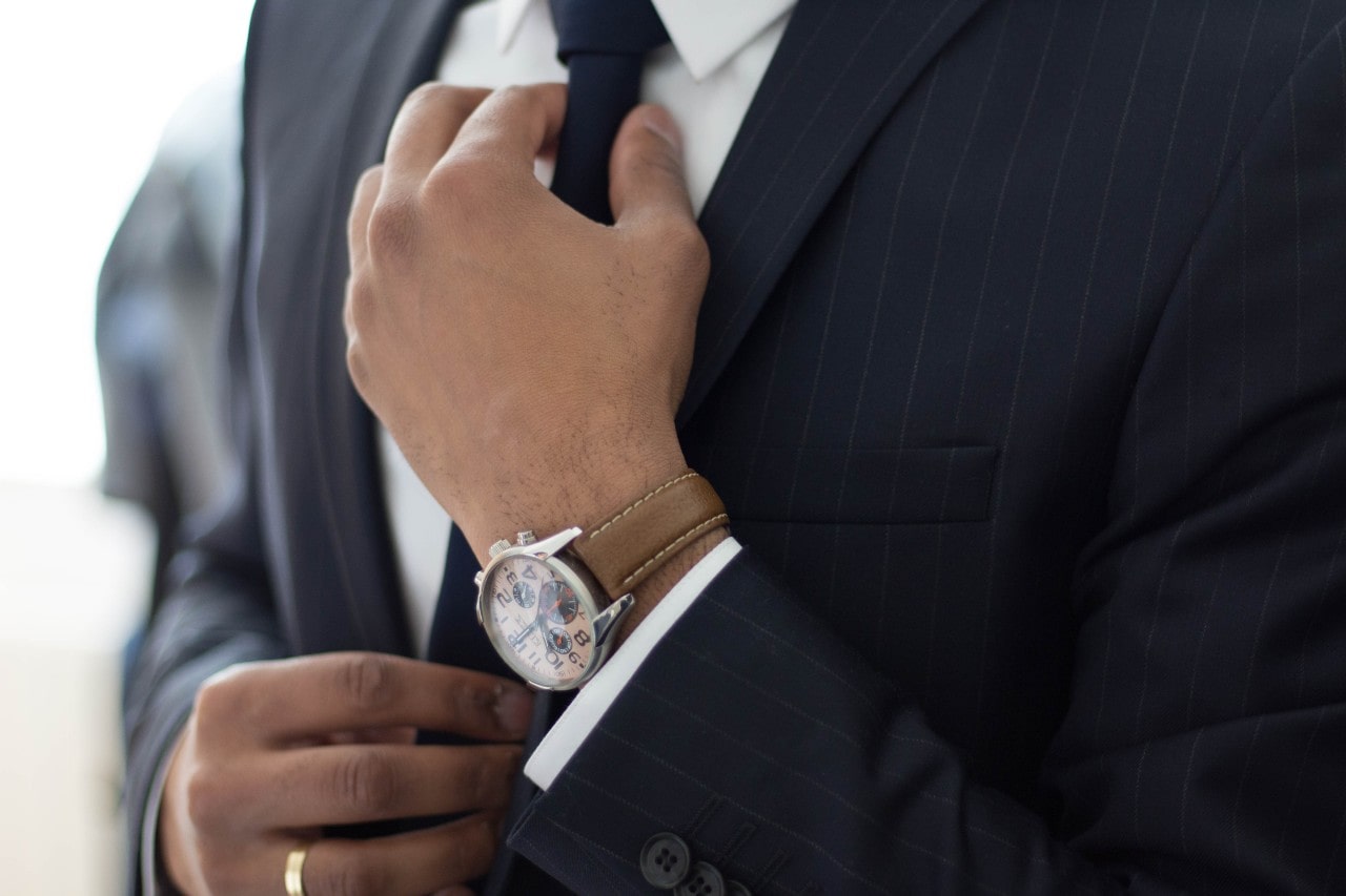 A close-up of a man in a grey striped suit fixing his tie, showcasing a brown leather wristwatch and a yellow gold wedding band.