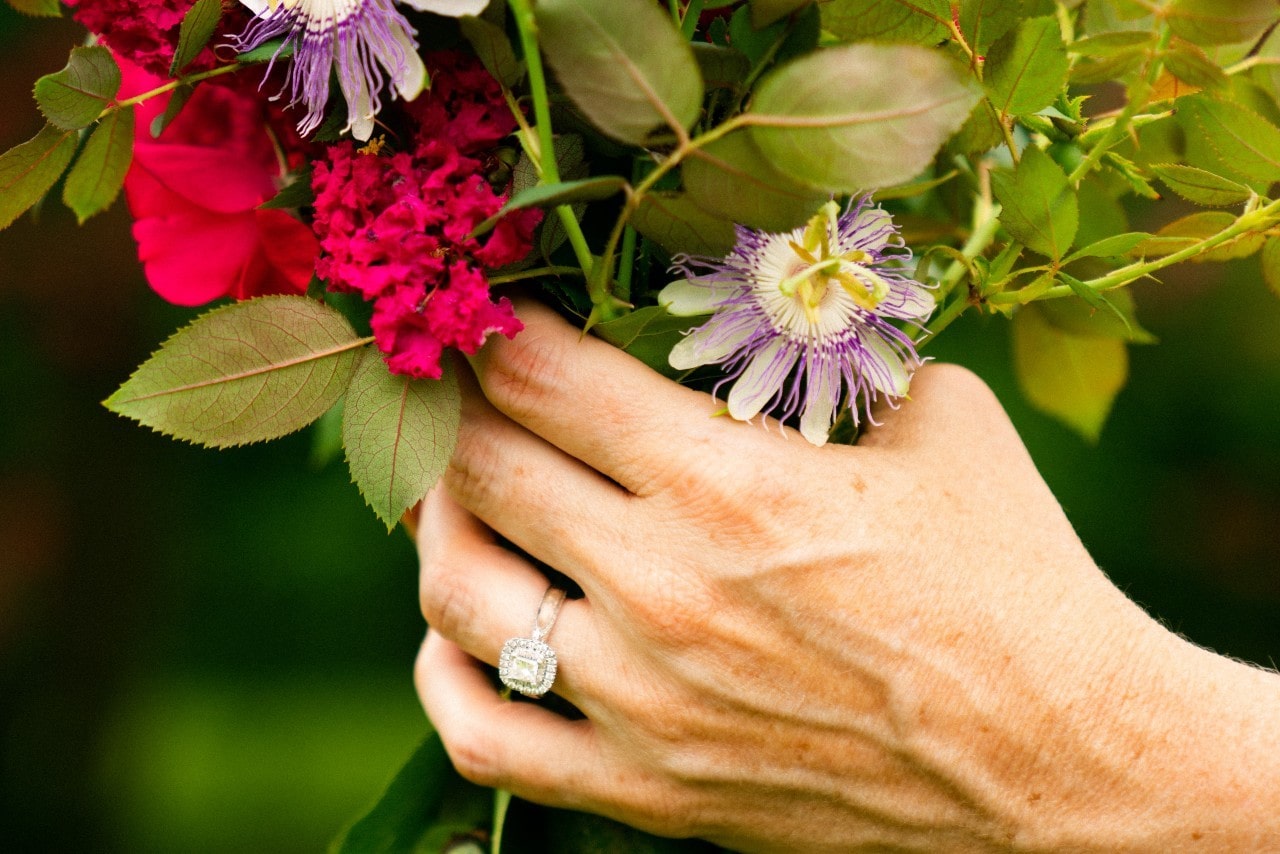 a woman’s hand holding a colorful bouquet of flowers and wearing a white gold halo engagement ring