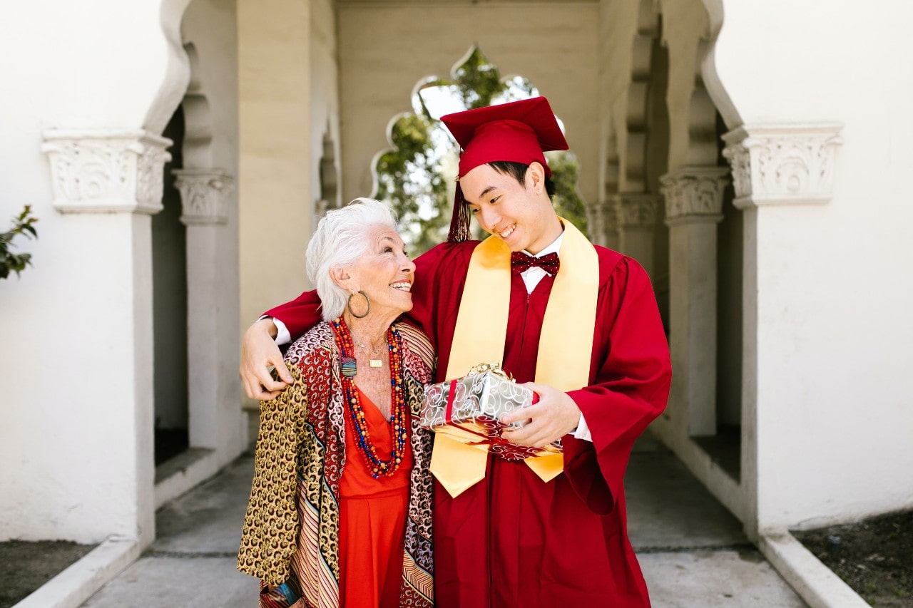 A graduate in a red gown hugs his grandmother, holding a present in the other hand.