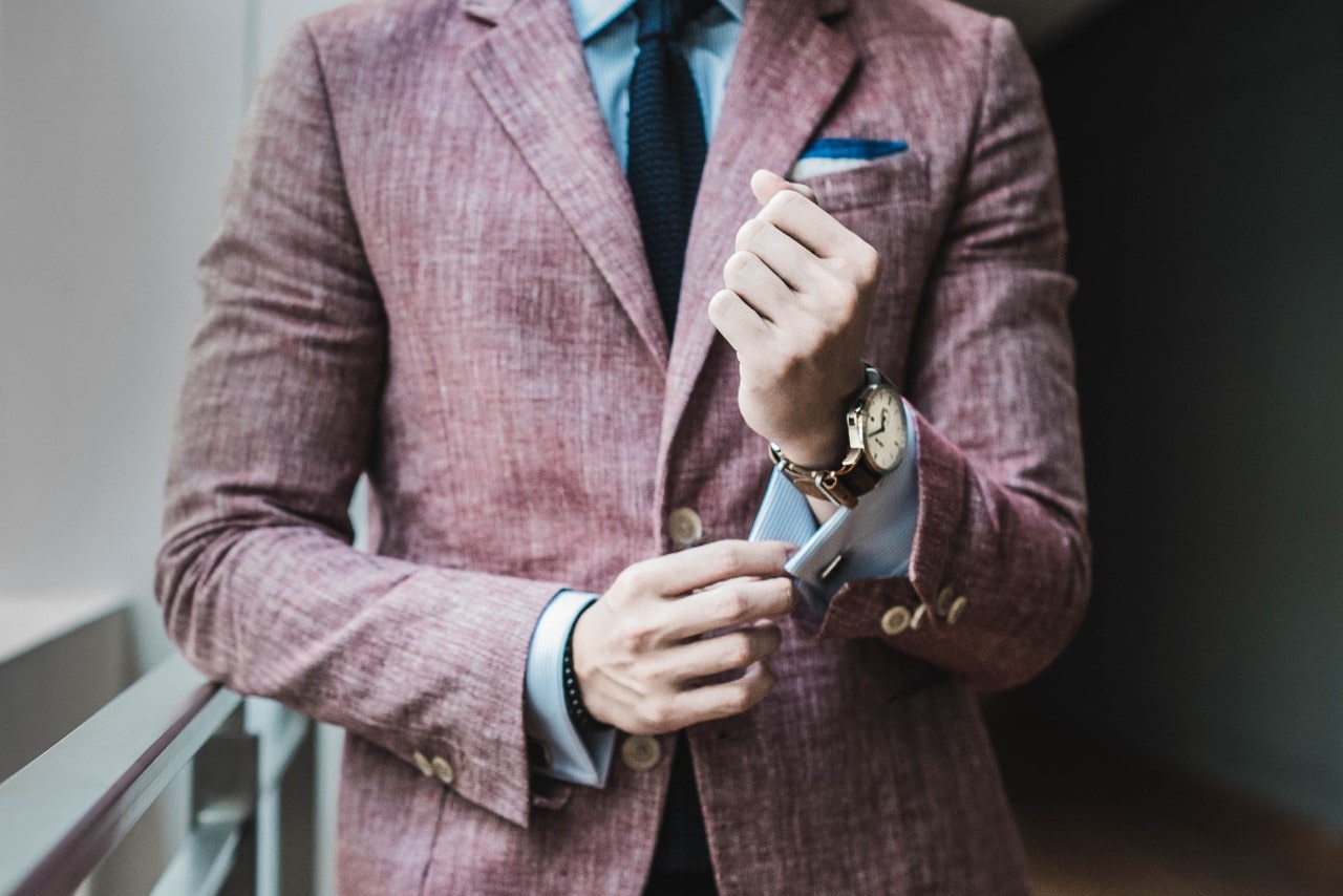 A man in a pink suit fixes his cufflinks, showcasing a leather strap watch.