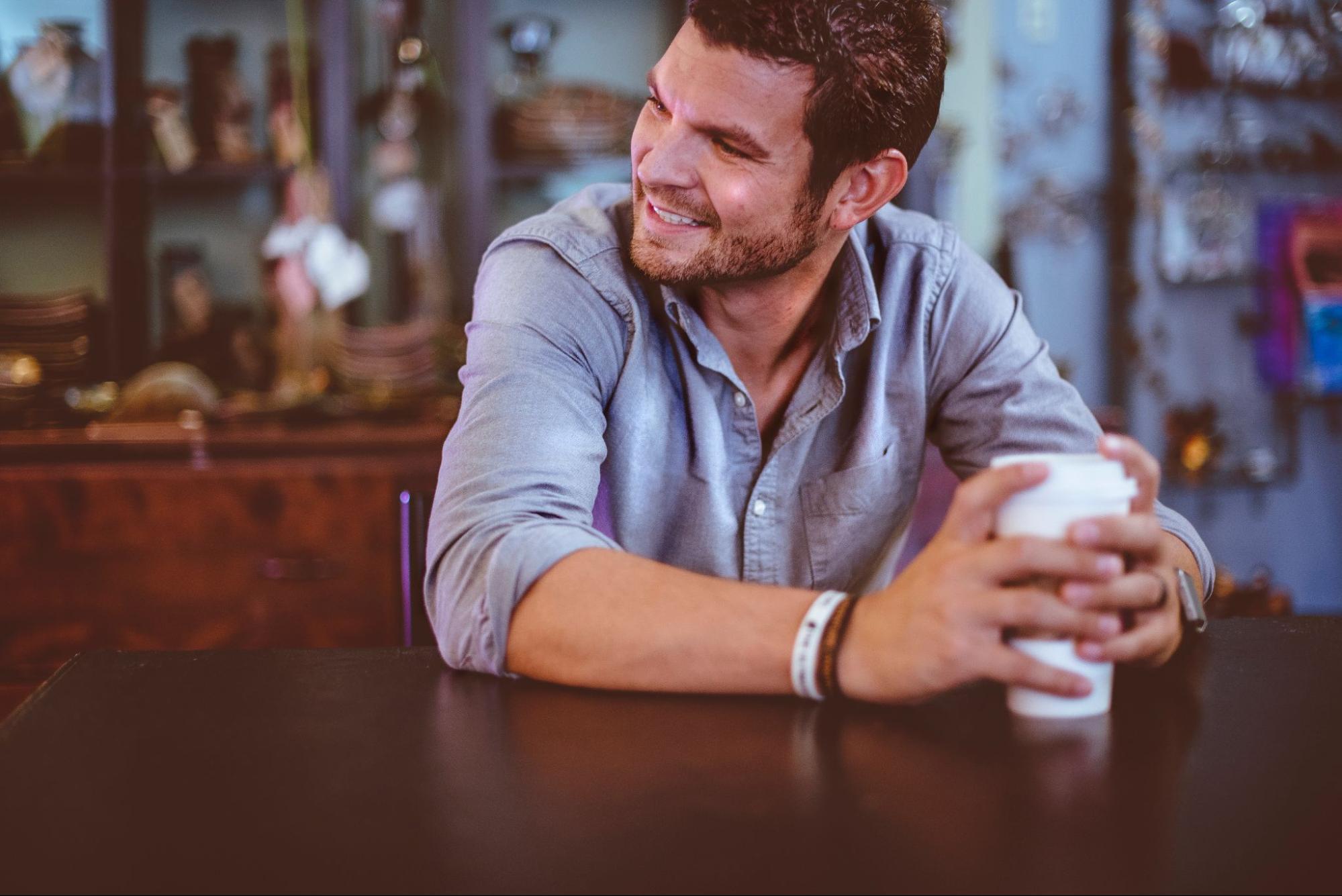 A man in a blue shirt sits at a table, holding a white cup and showcasing several rubber bracelets.