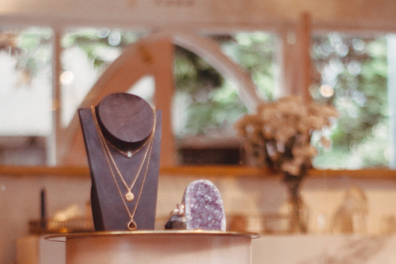 A display of elegant gold necklaces on a dark stand in a softly lit jewelry boutique, with blurred greenery and flowers in the background.