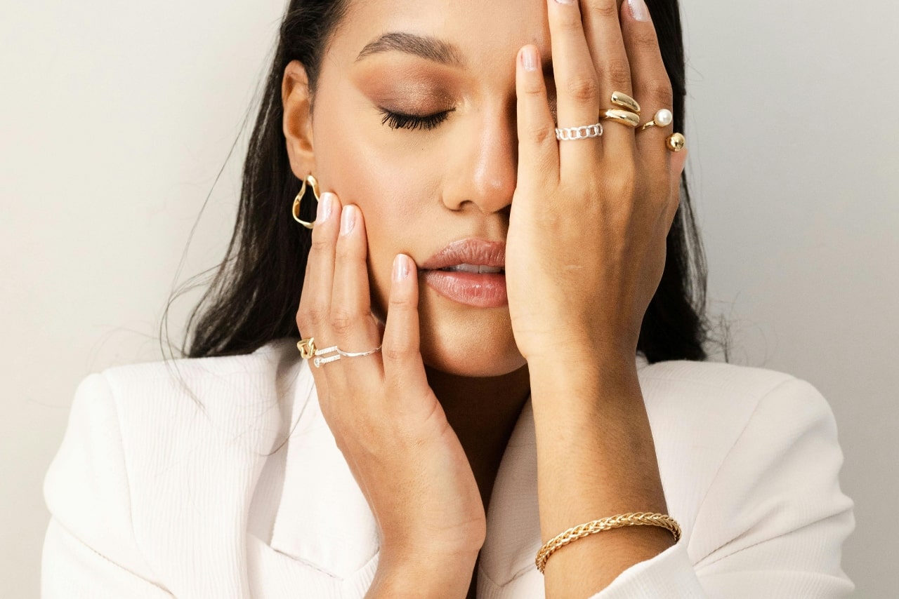 Woman in a white blazer, eyes closed, hand partially covering her face, flaunting gold and silver rings
