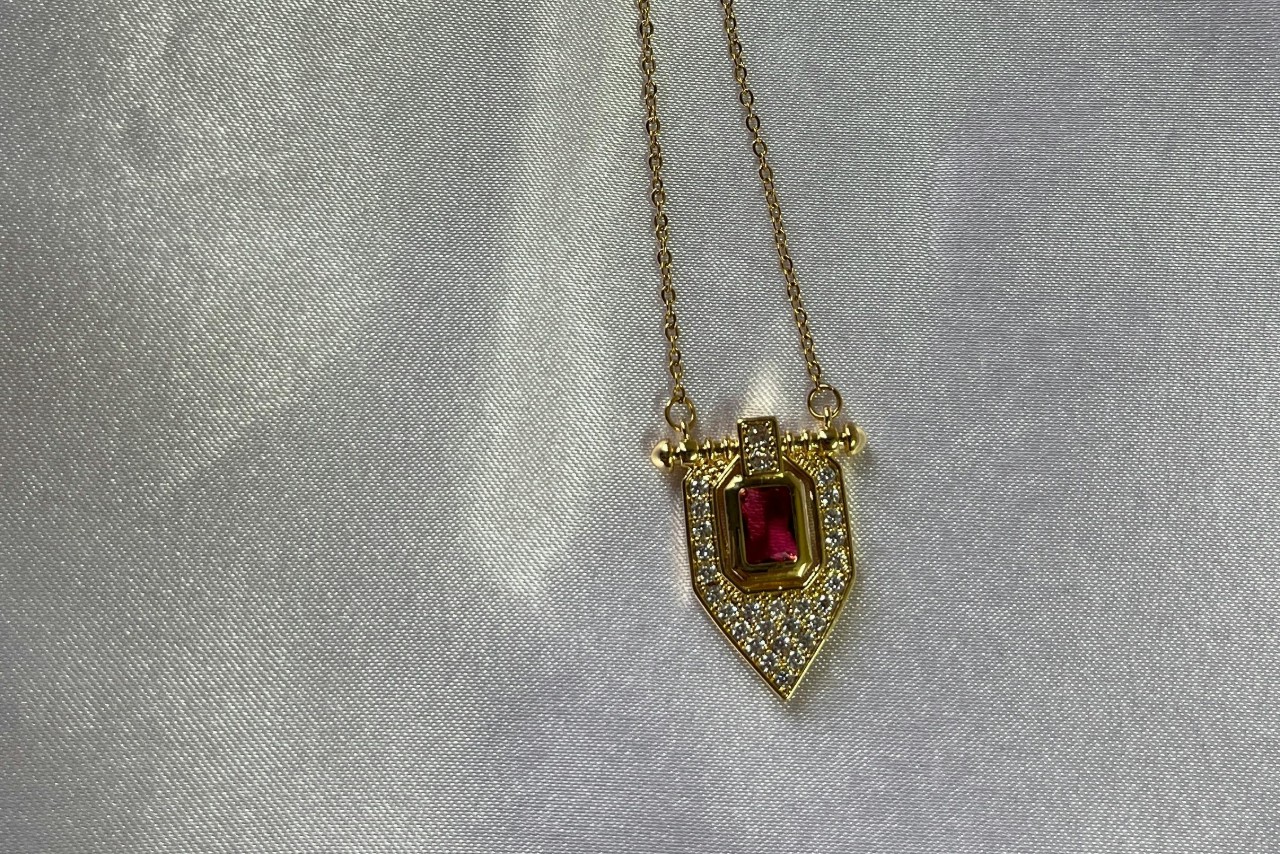A yellow gold geometric ruby pendant necklace with diamond accents on a gray surface.