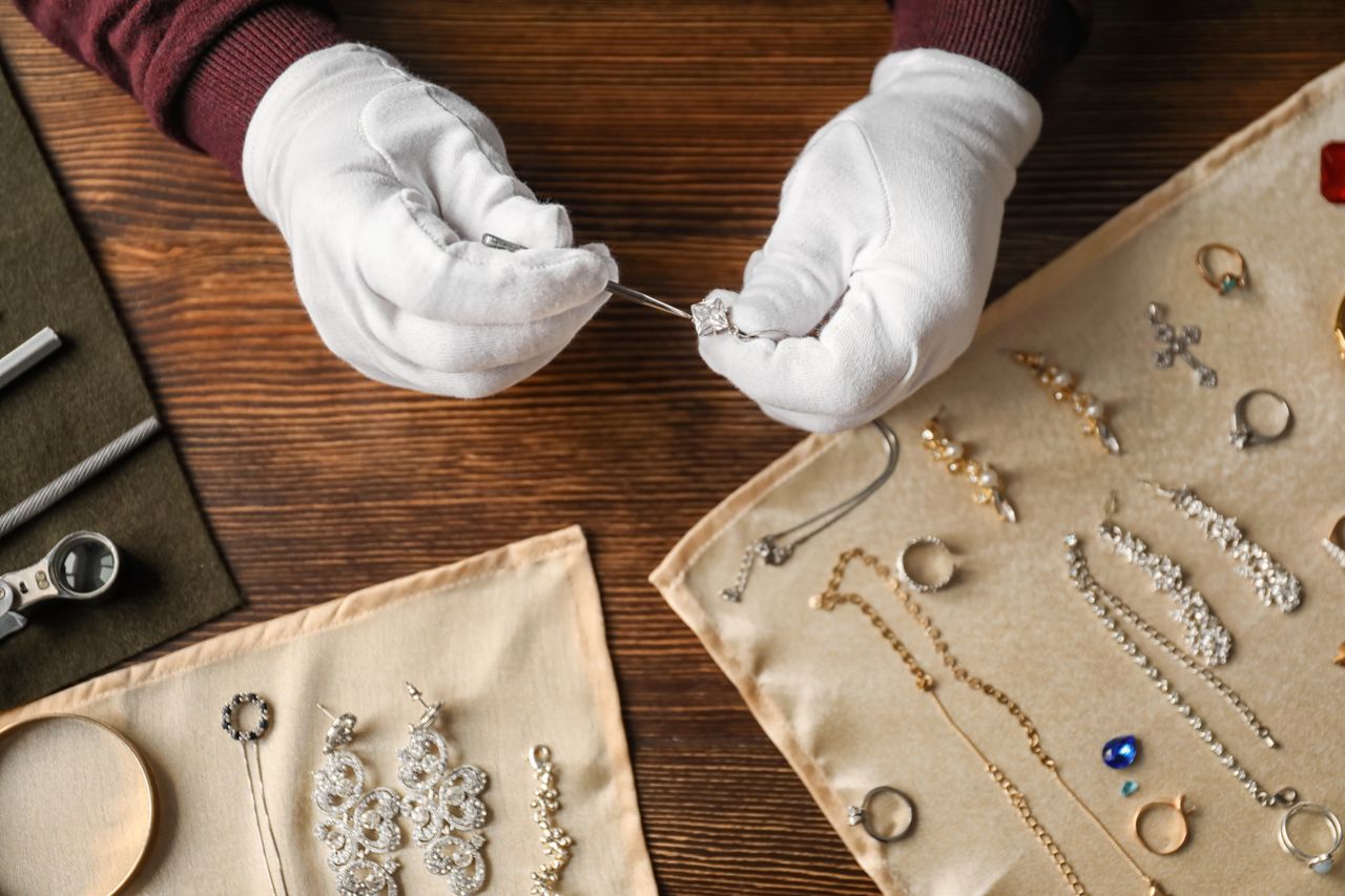 Close-up of a jeweler's gloved hands using tweezers to fix a diamond pendant, with several pieces of jewelry laid out on beige clothes on a wooden table.