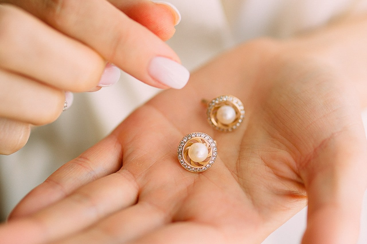A close-up of a hand holding a pair of gold earrings featuring central pearls surrounded by a halo of small diamonds. A close-up of a hand holding a pair of gold earrings featuring central pearls surrounded by a halo of small diamonds.