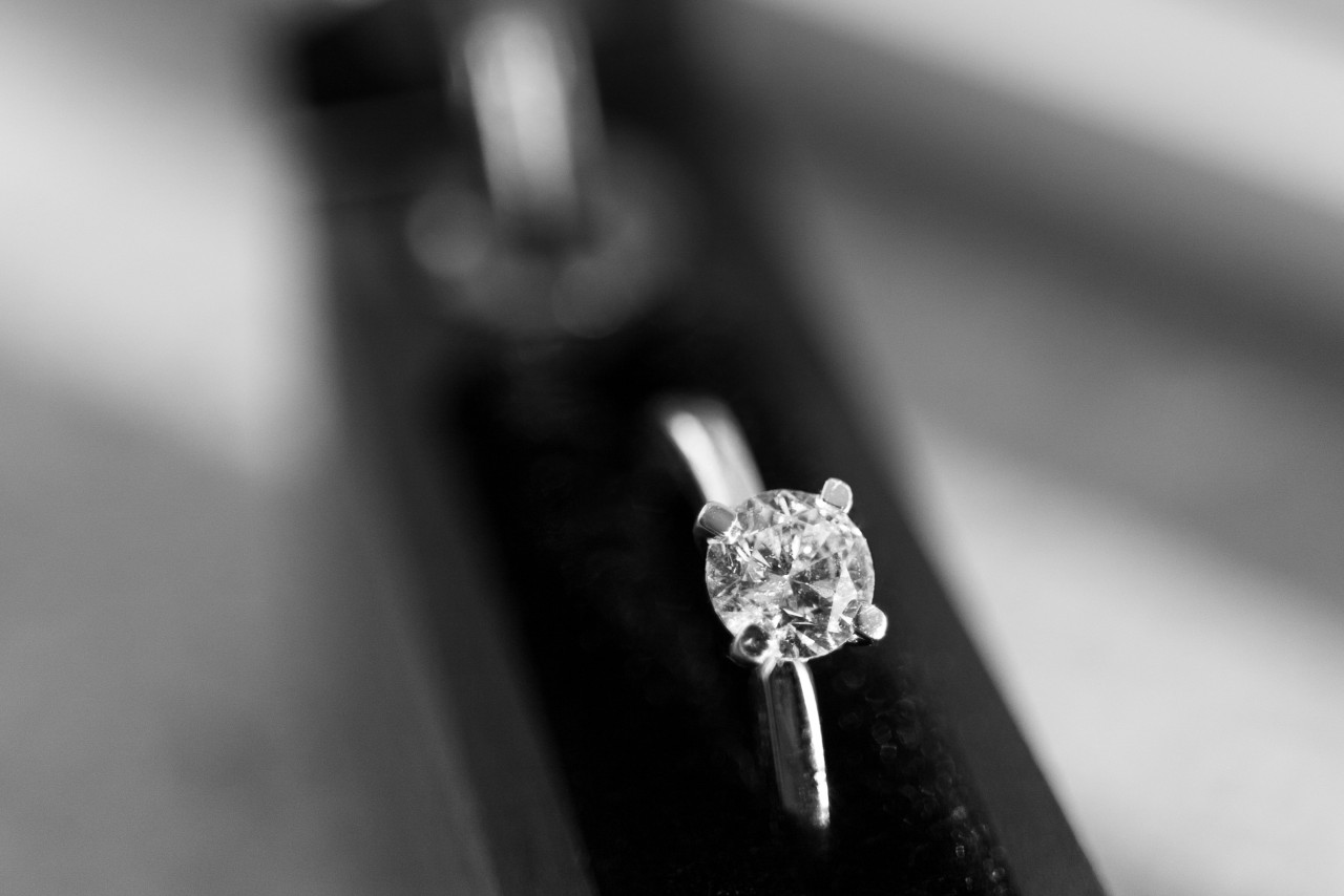 A black and white close-up of a simple but elegant solitaire engagement ring with a round center stone displayed upright.