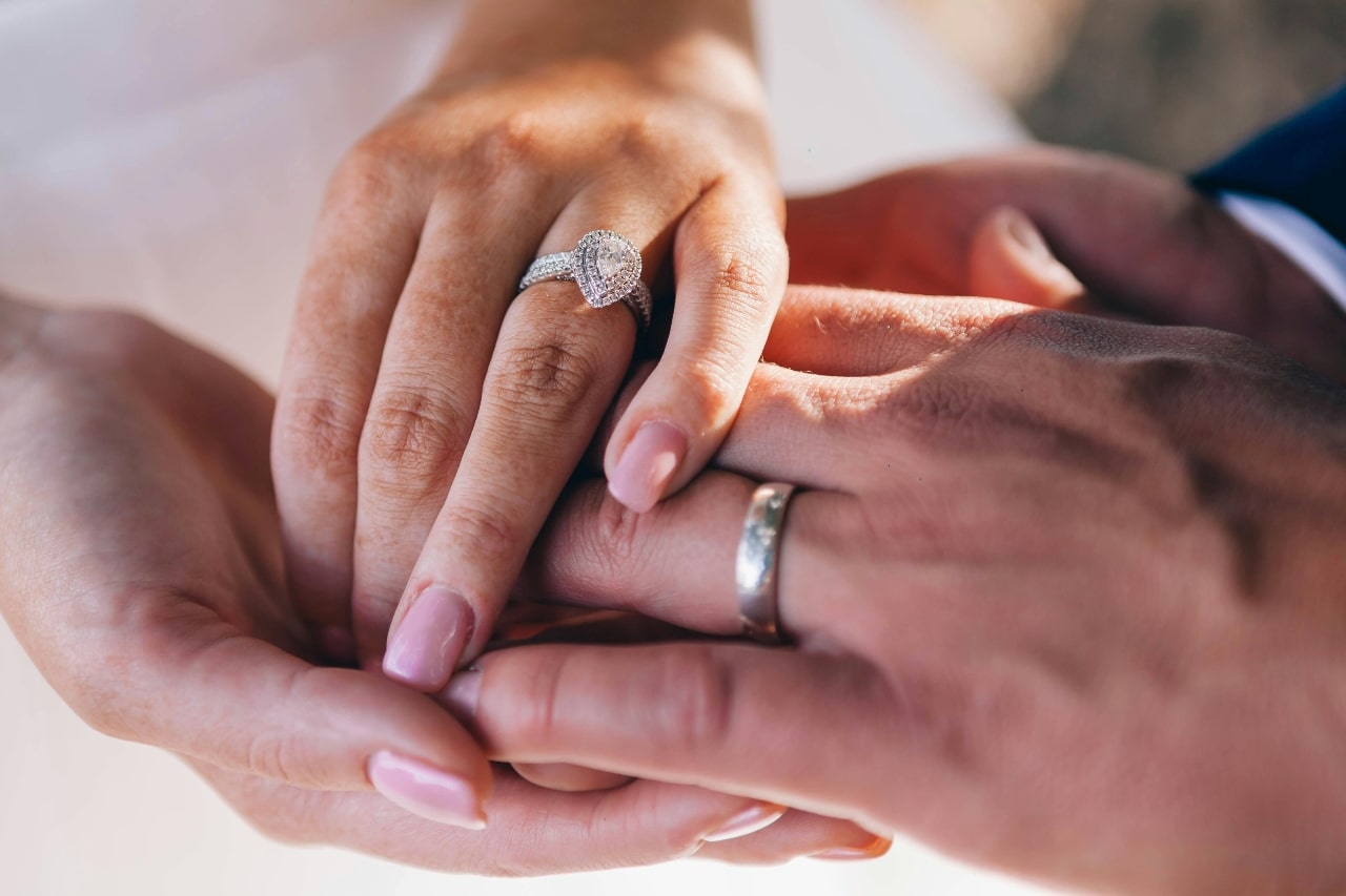 A close-up of a couple holding hands, with emphasis on the man’s wedding band and the woman’s ornate engagement ring.