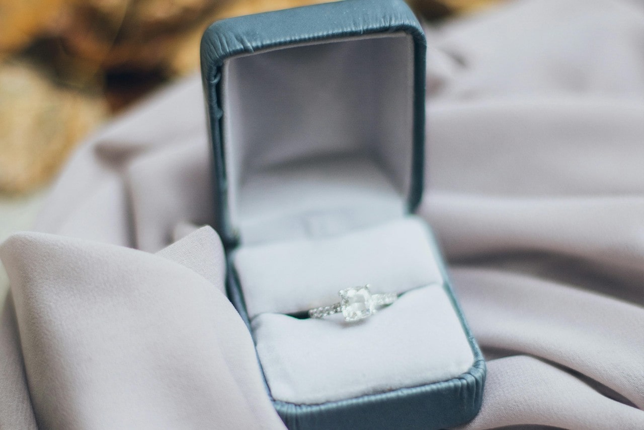 A close-up of a side stone engagement ring with a cushion cut center diamond displayed in-box atop textured taupe fabric.