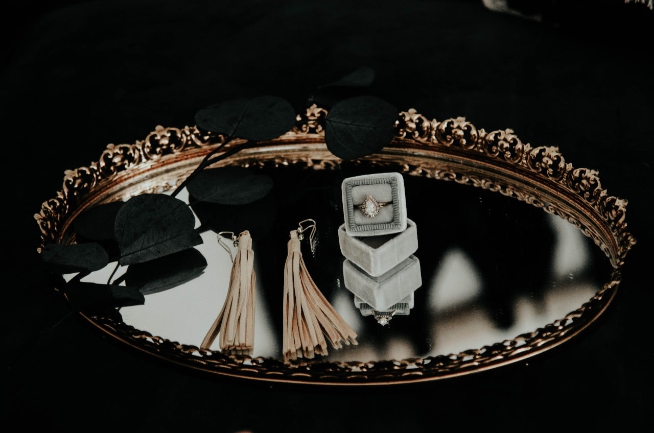 A ring box, fringe earrings, and black flowers sit in a golden mirror tray.