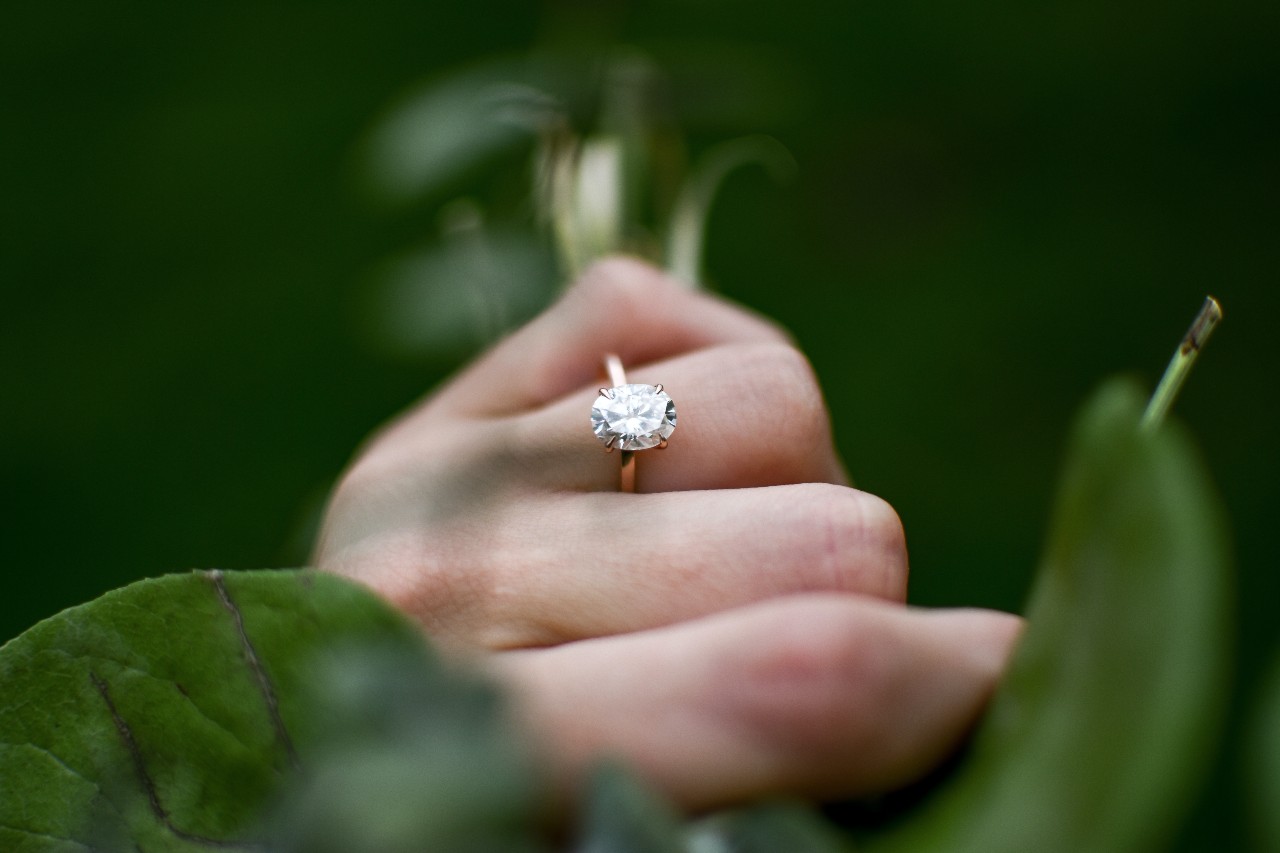 a hand wearing an oval cut engagement ring and surrounded by greenery
