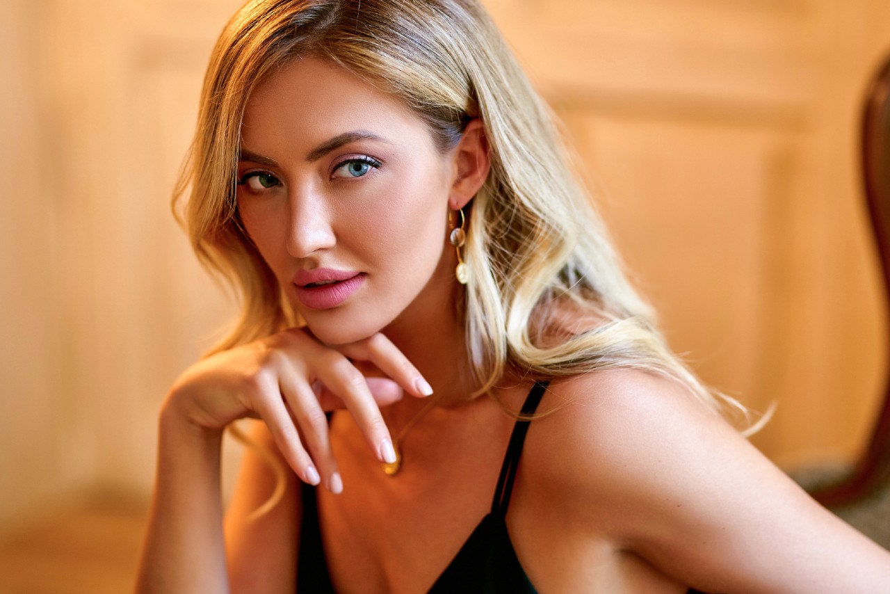 A blonde woman wearing a black spaghetti strap top and gold dangle earrings, posing against a blurred warm-toned background.