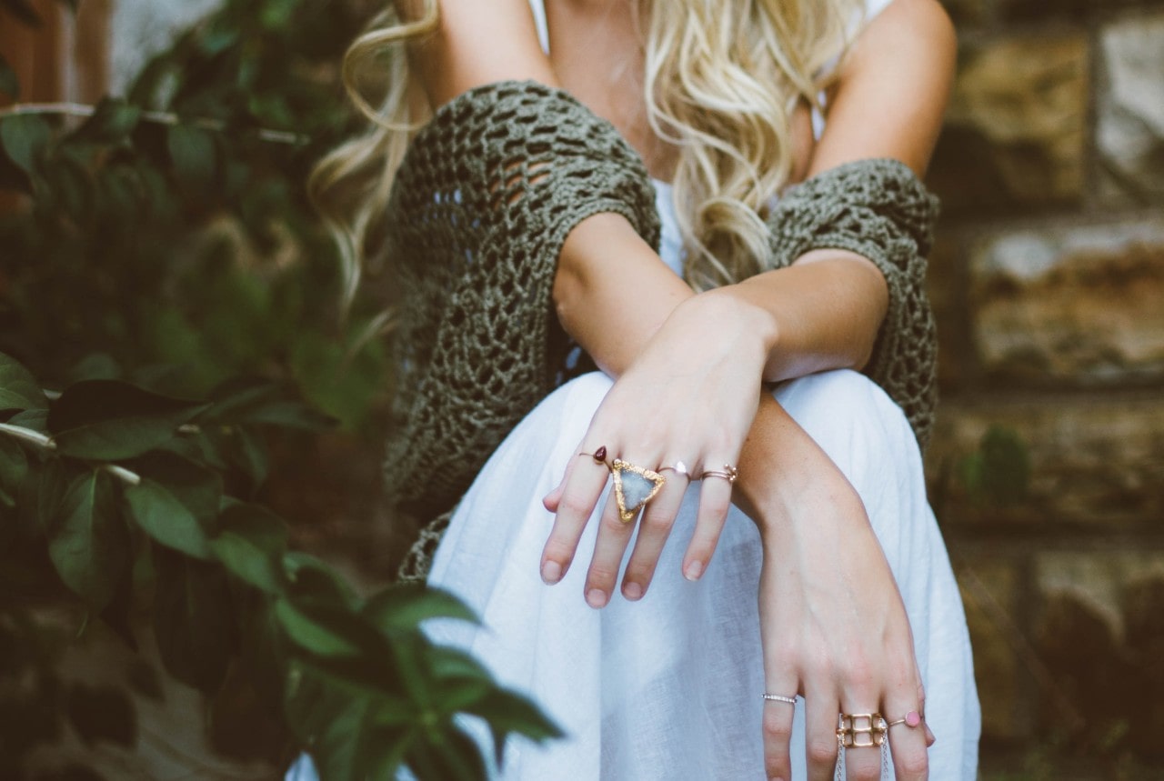A blonde woman wearing a flowy white dress and a green crocheted shawl in front of a brick wall and greenery, with gold rings on her fingers.