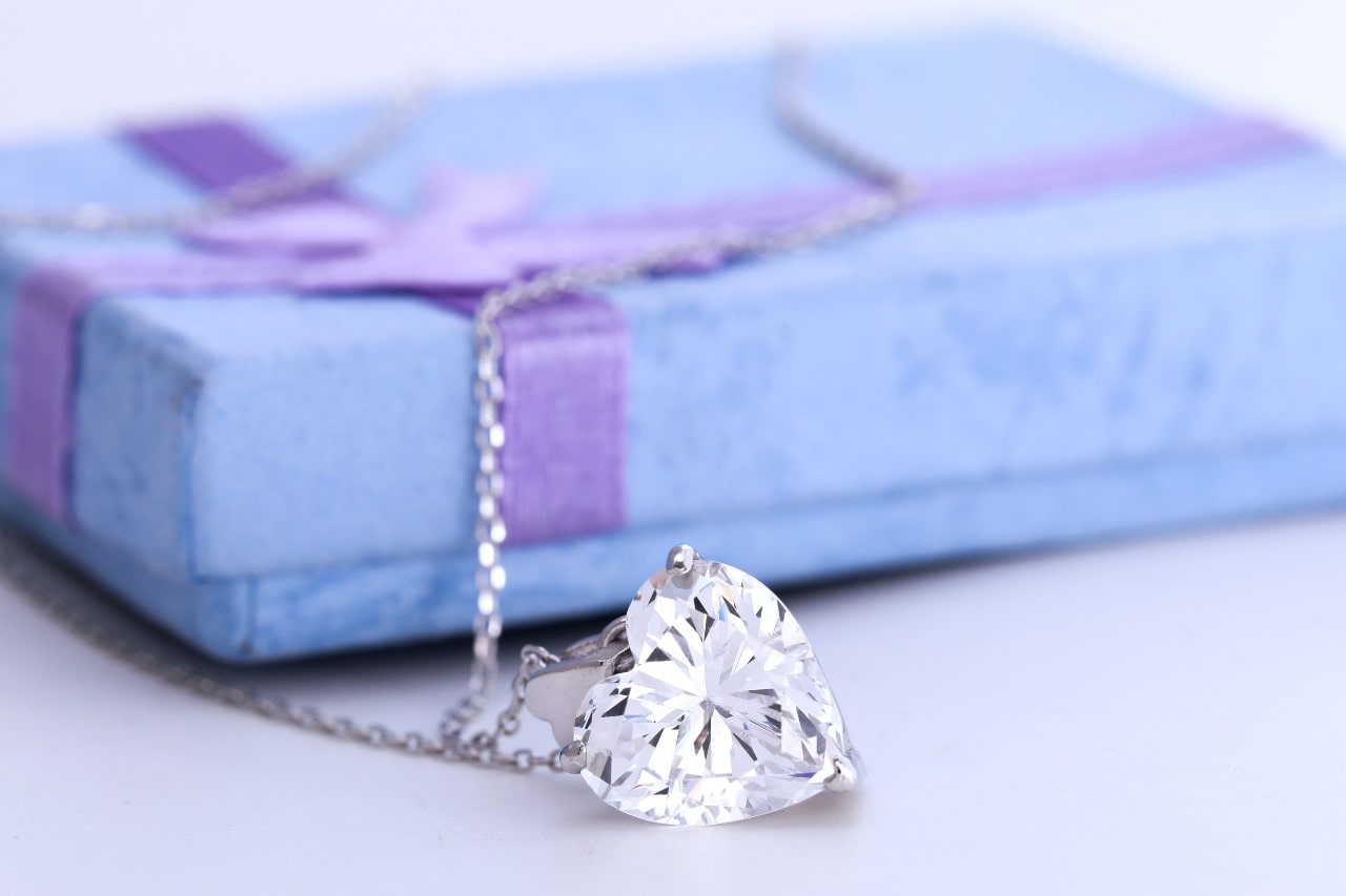 A close-up of a necklace with a heart-shaped diamond pendant displayed on a blue gift box with purple ribbon. A close-up of a necklace with a heart-shaped diamond pendant displayed on a blue gift box with purple ribbon.