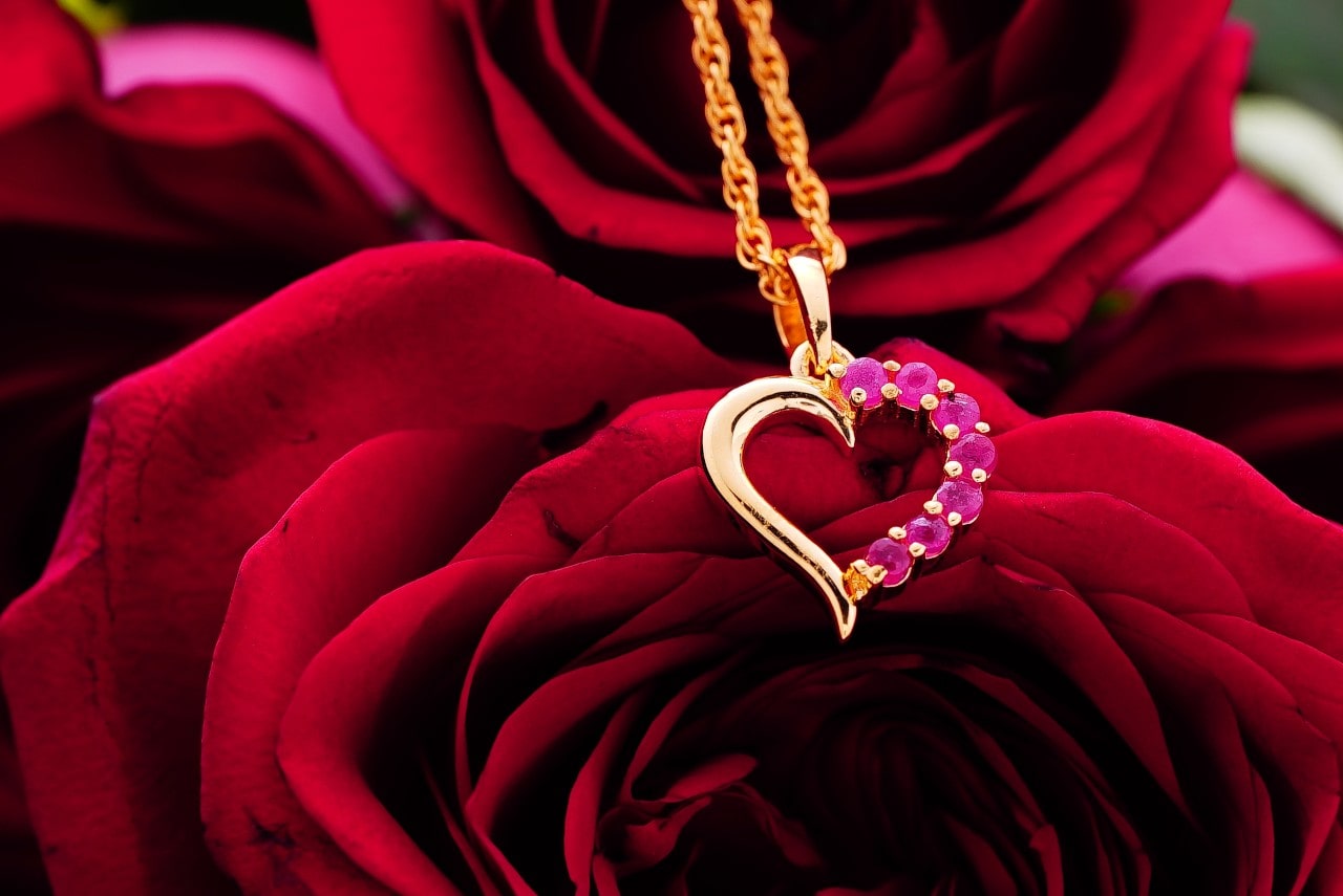 A yellow gold heart pendant that is encrusted with rubies on one half lay on red roses A yellow gold heart pendant that is encrusted with rubies on one half lay on red roses