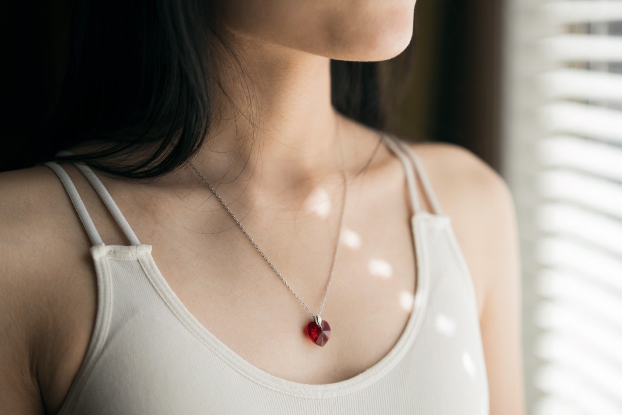 A close-up of a woman wearing a red heart pendant necklace near a window with soft light.