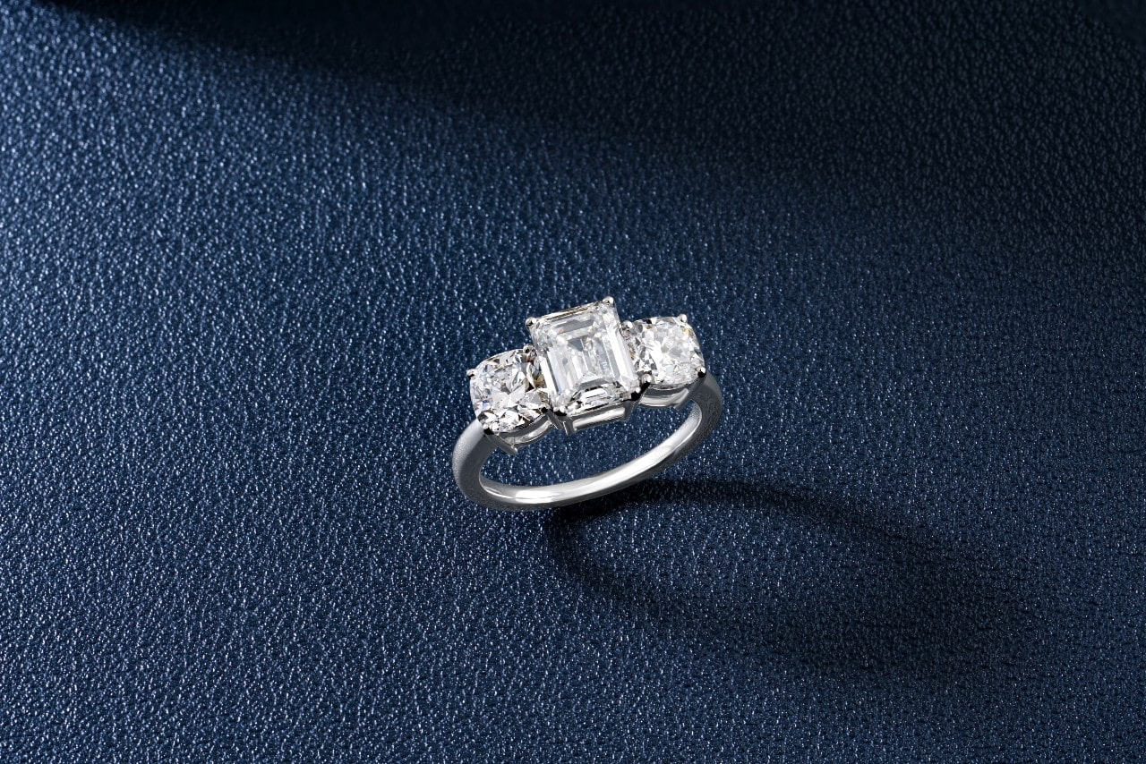 A platinum three stone diamond engagement ring sits on a textured black surface.