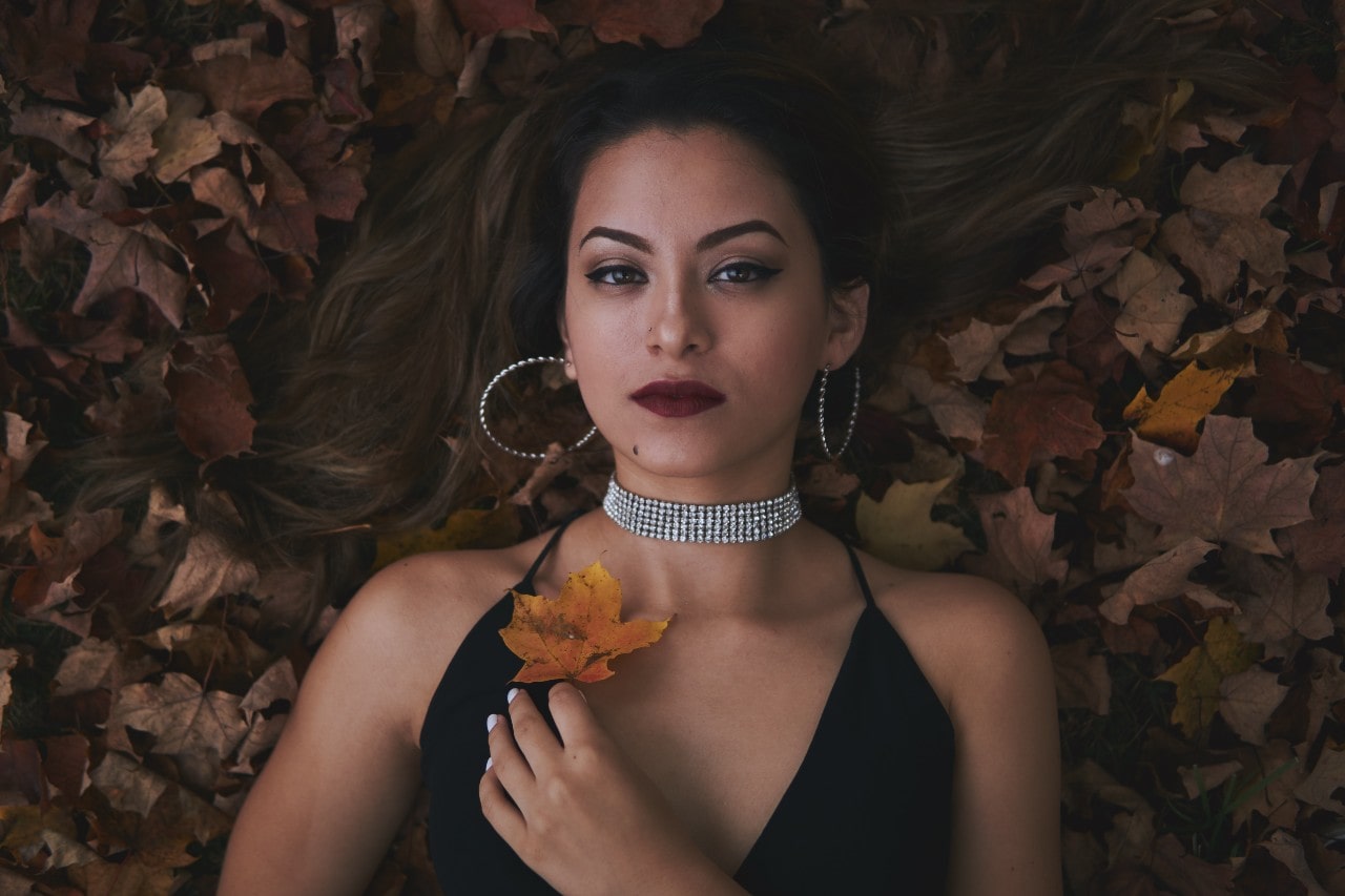 A woman in a formal dress lays down in a pile of leaves while wearing a diamond choker.