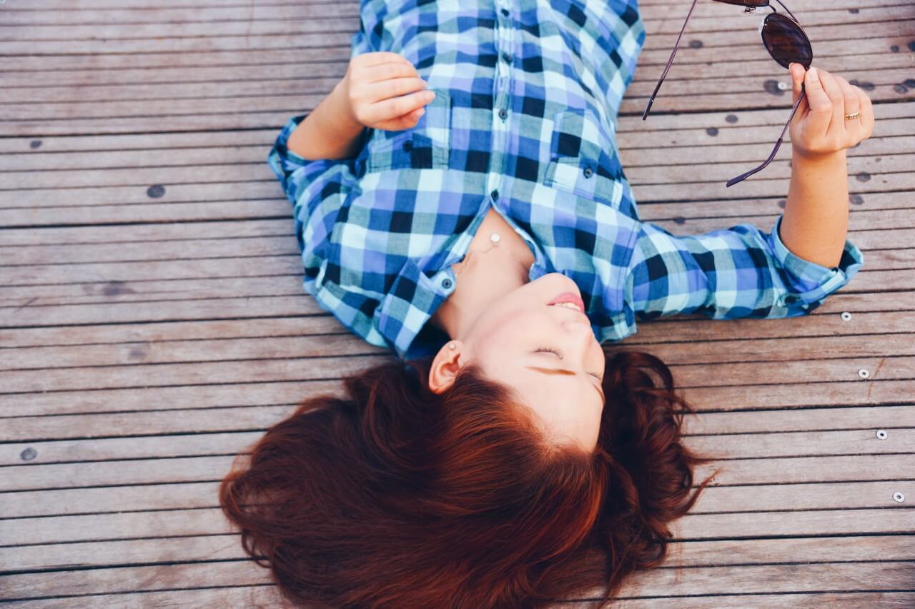 A woman lays down on a dock while holding sunglasses and wearing a diamond necklace.