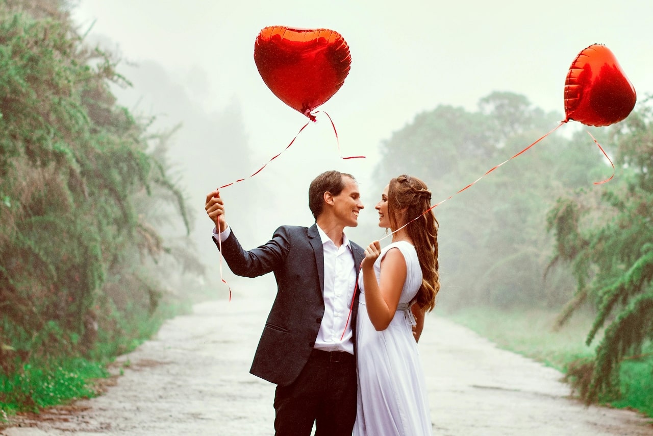 A man and a woman embrace each other, while holding red heart shaped balloons against a rainy forest.