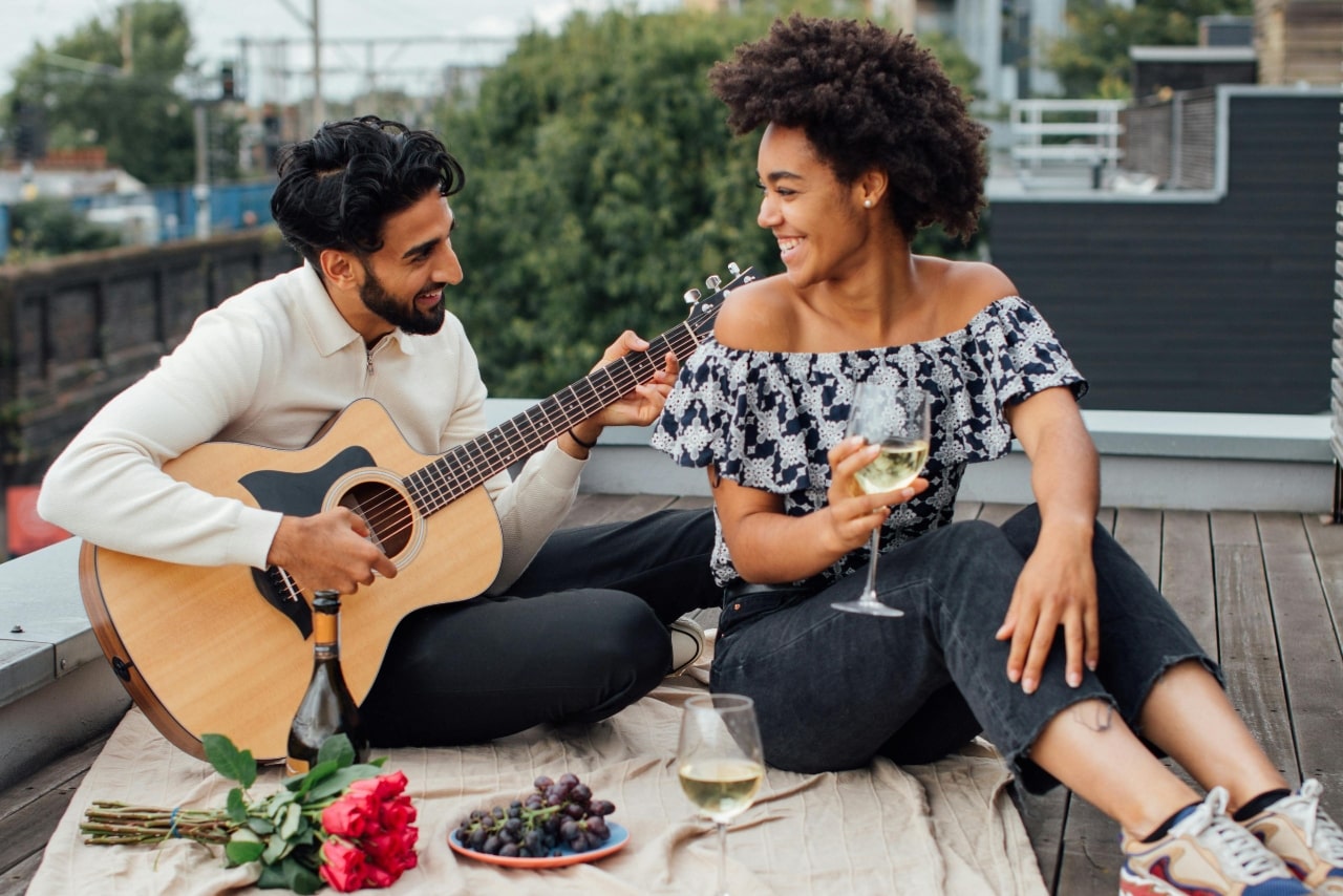 A couple engages in a relaxed rooftop touchpoint, with the man strumming a guitar while the woman enjoys a glass of white wine amid a setup featuring roses, grapes, and a bottle of wine