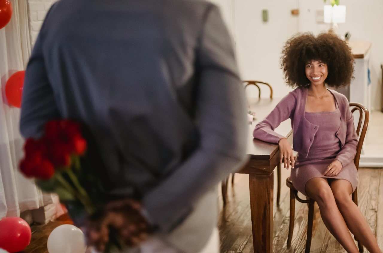 A man hides a bouquet of roses behind his back as he approaches a smiling woman seated at a table in a warmly decorated, festive room