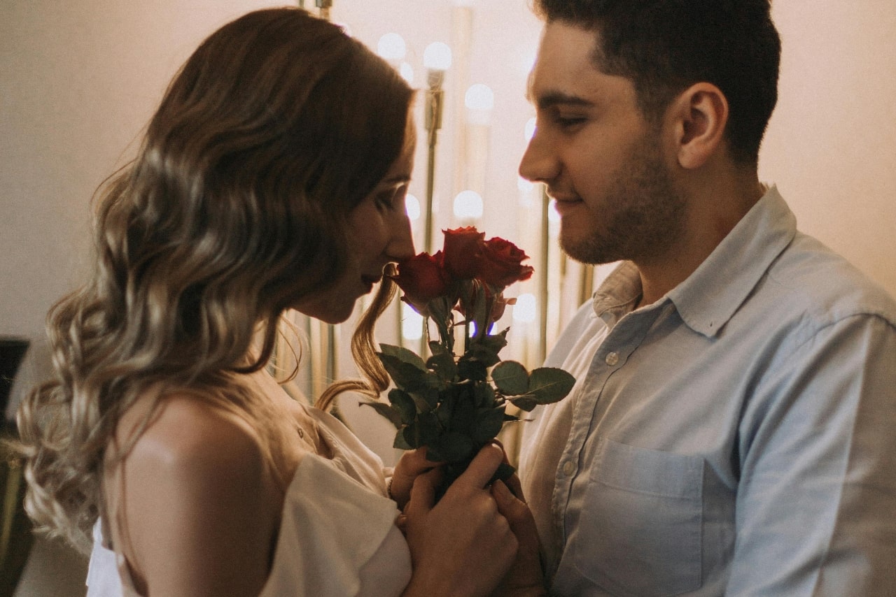 A close up of a woman with wavy hair and a man in a shirt embracing, while the woman smells red roses.