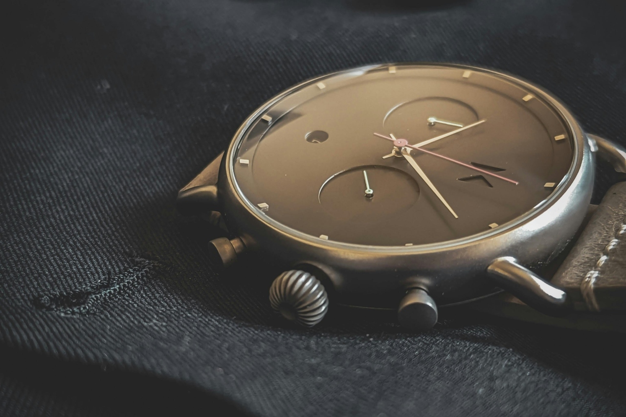 Close-up of a modern wristwatch with a minimalist black dial, silver watch hands, on a textured dark fabric background.