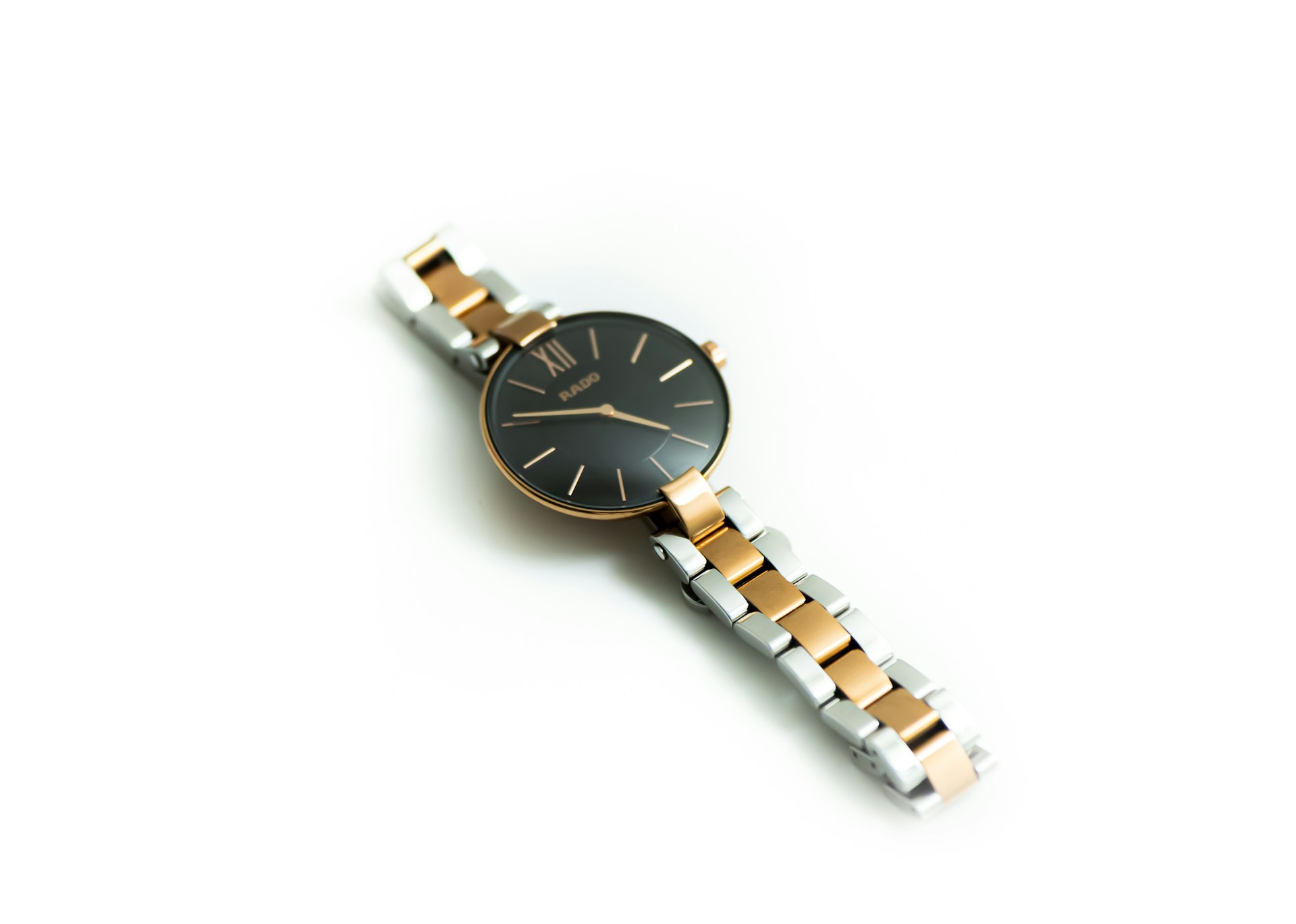 A wristwatch with a silver and rose gold metal band on a white surface, with the black face featuring minimalist hour markers and hands