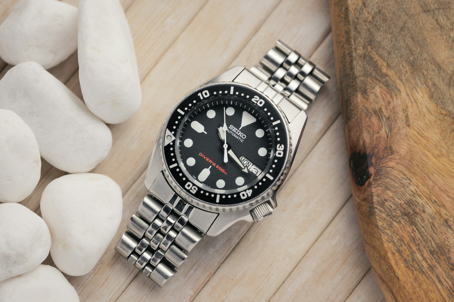 A silver Seiko watch with a black face and white markers rests on a wooden surface, surrounded by smooth white stones