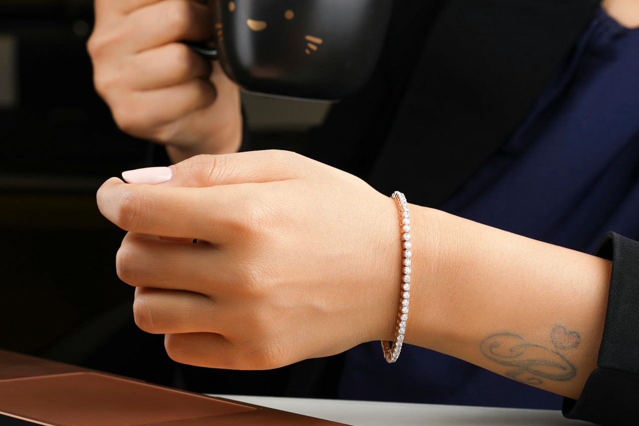 A person with a heart and initial wrist tattoo holds a mug while wearing a yellow gold diamond tennis bracelet. A person with a heart and initial wrist tattoo holds a mug while wearing a yellow gold diamond tennis bracelet.