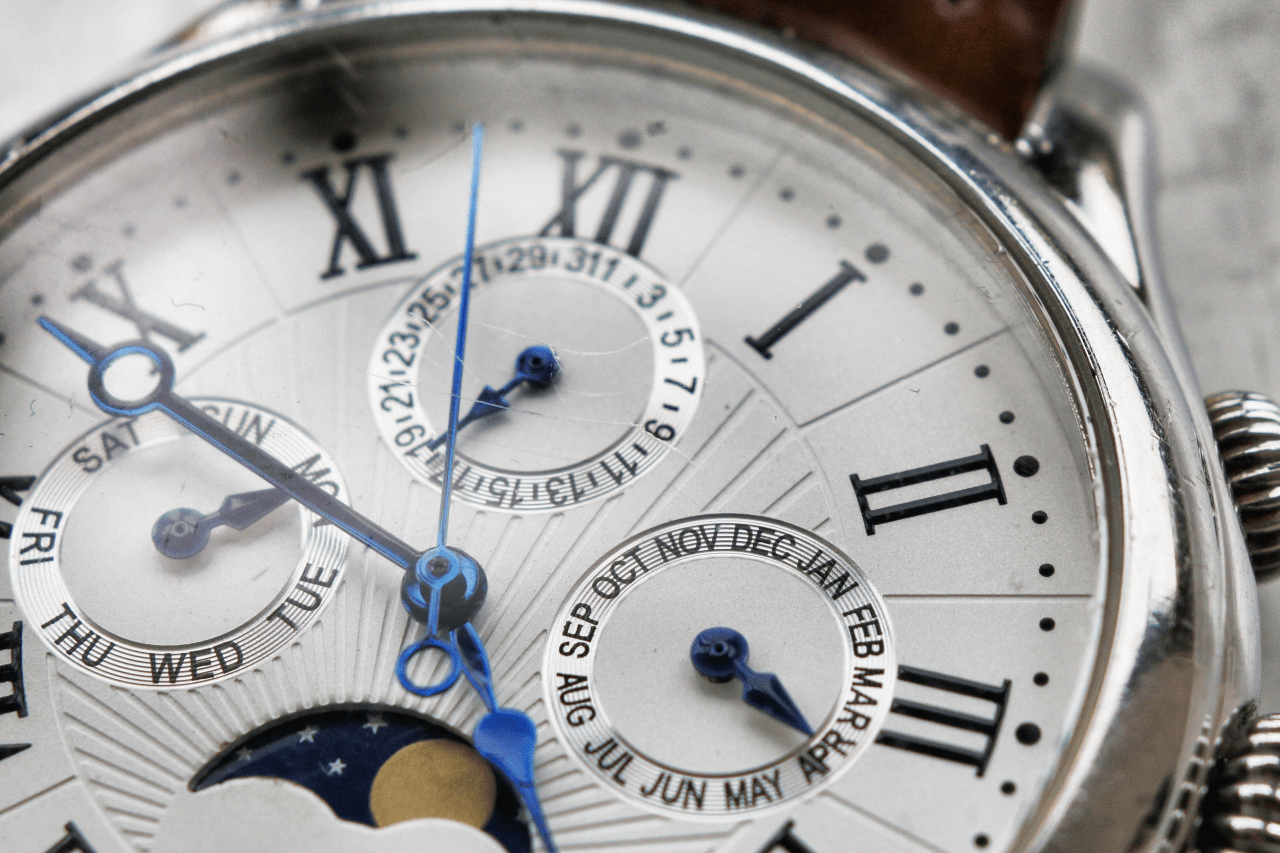 A macro shot of a wristwatch face with Roman numerals, multiple subdials, and blue hands. A macro shot of a wristwatch face with Roman numerals, multiple subdials, and blue hands.